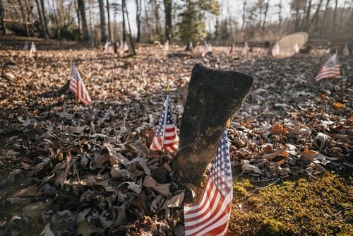 A weathered gravestone marked with small American flags among fallen autumn leaves
